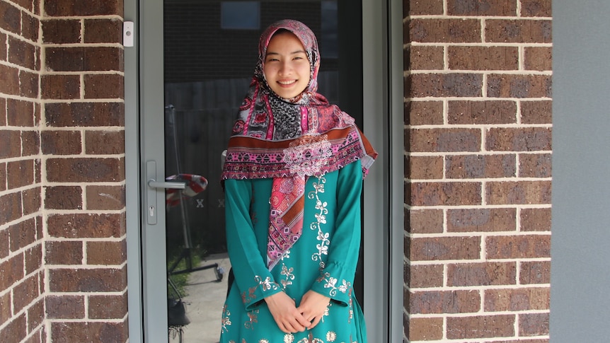 A young woman in a bright pink patterned hijab stands outside the front door of her brown brick home.