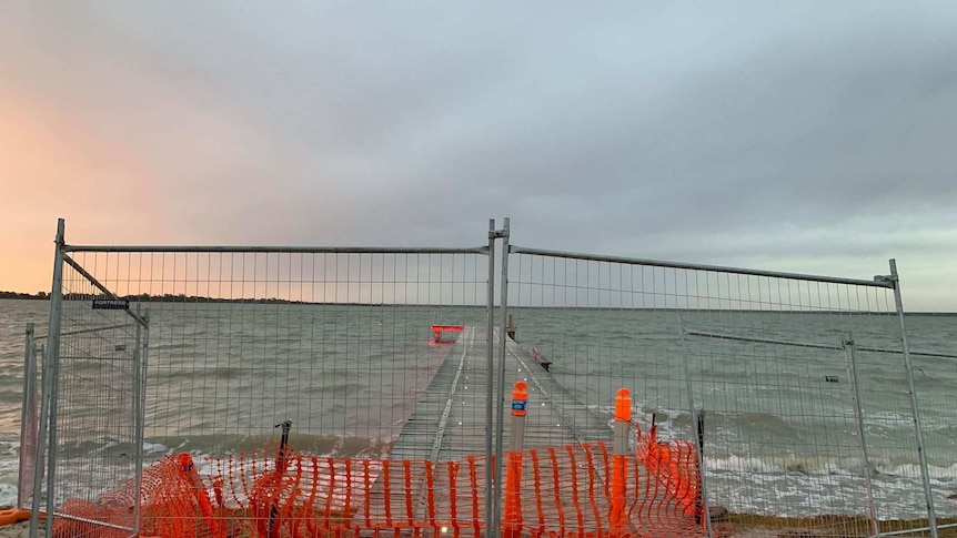 A fence is standing in front of a jetty, blocking access. There is water all around and the sun is setting.
