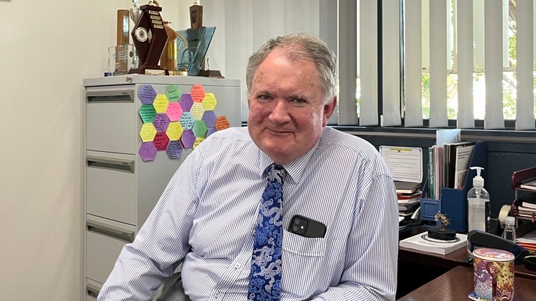 older man sits behind desk in striped shirt and partnered tie 