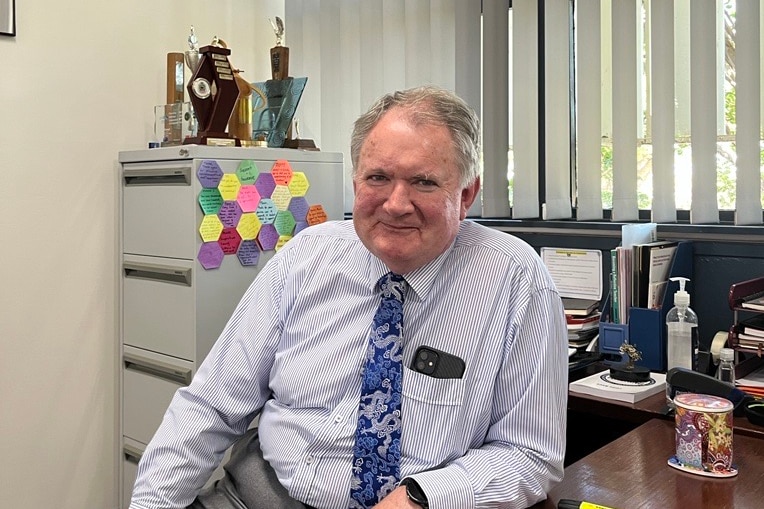 older man sits behind desk in striped shirt and partnered tie 