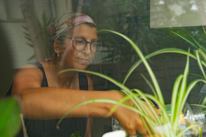A woman wearing a bright headscarf waters plants, the photo is taken through a window.