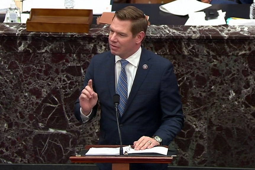A middle-aged man in a dark suit, white shirt and blue tie stands at lectern and raises his finger.