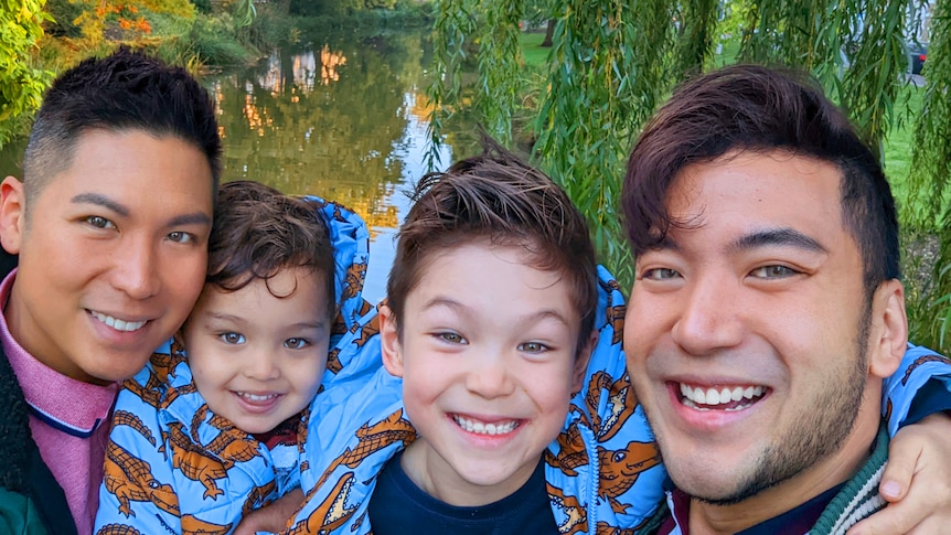 Two dads and two boys pose together with arms around one another smiling, against a forest backdrop.