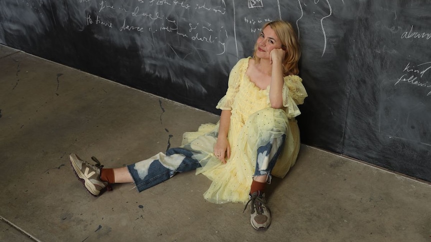 Jazz sits on a concrete floor and smiles up at the camera. They are wearing a yellow tulle dress, distressed jeans and trainers
