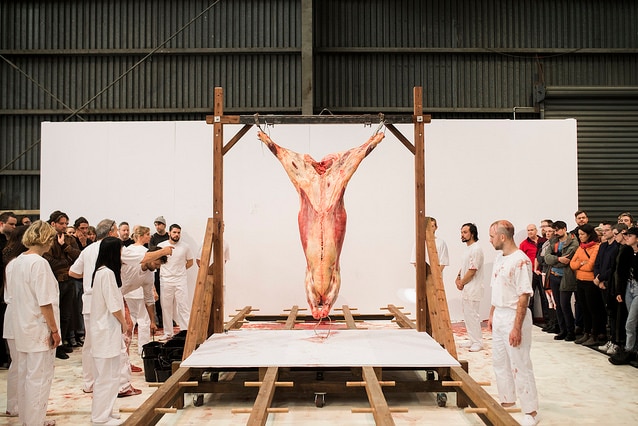 A bull carcass is held aloft in a scaffold, watched by people.