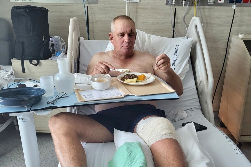 A man with a bandage wrapped knee eating dinner in a hospital bed.