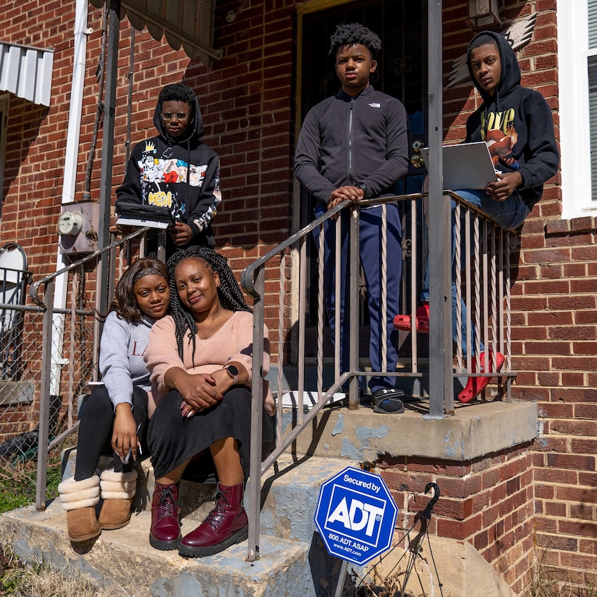 A woman and a teen girl sit on a stoop with three teen boys standing behind them