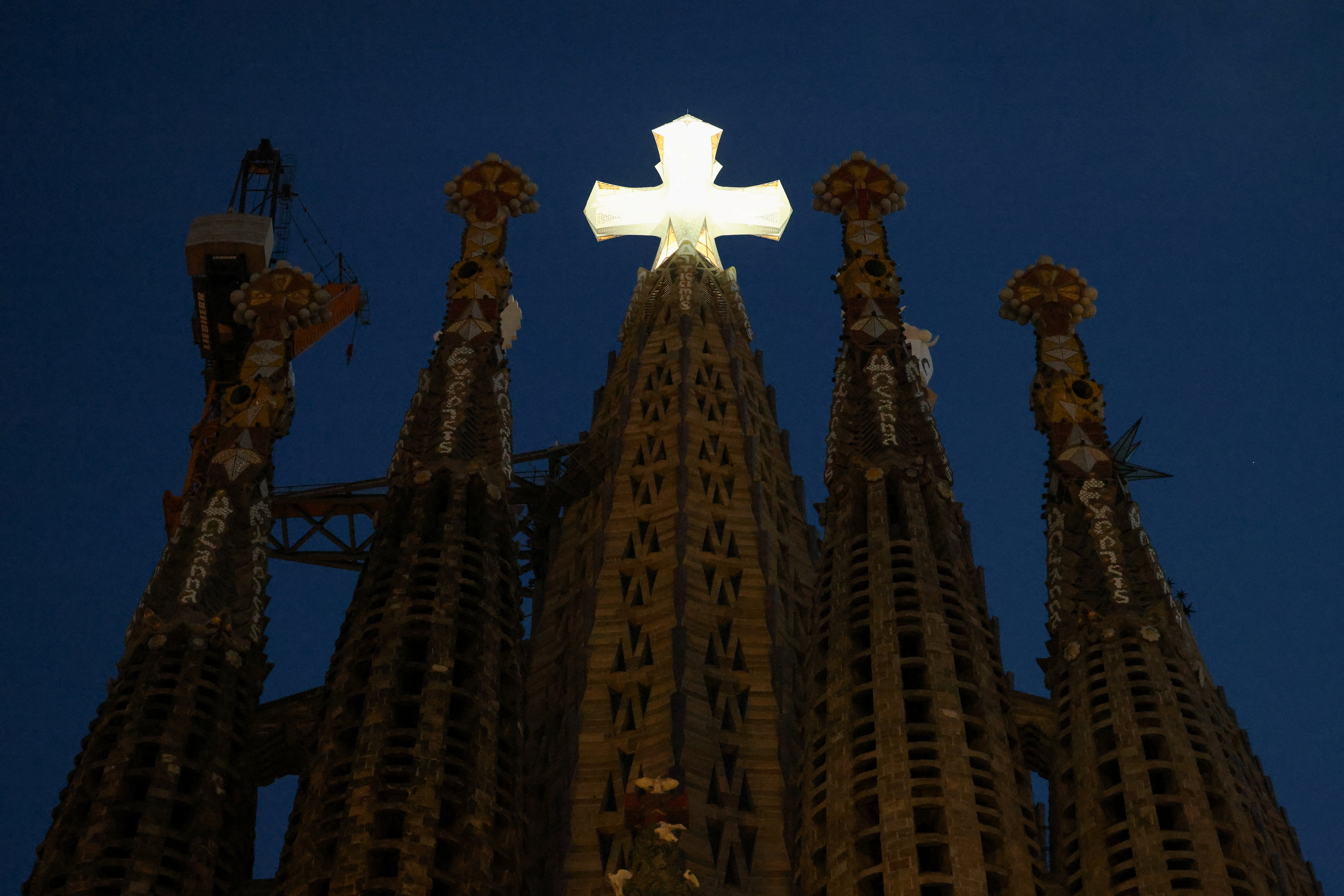 Five Sagrada Familia pylons at night, with a cross atop the central tower illuminated in bright white light.