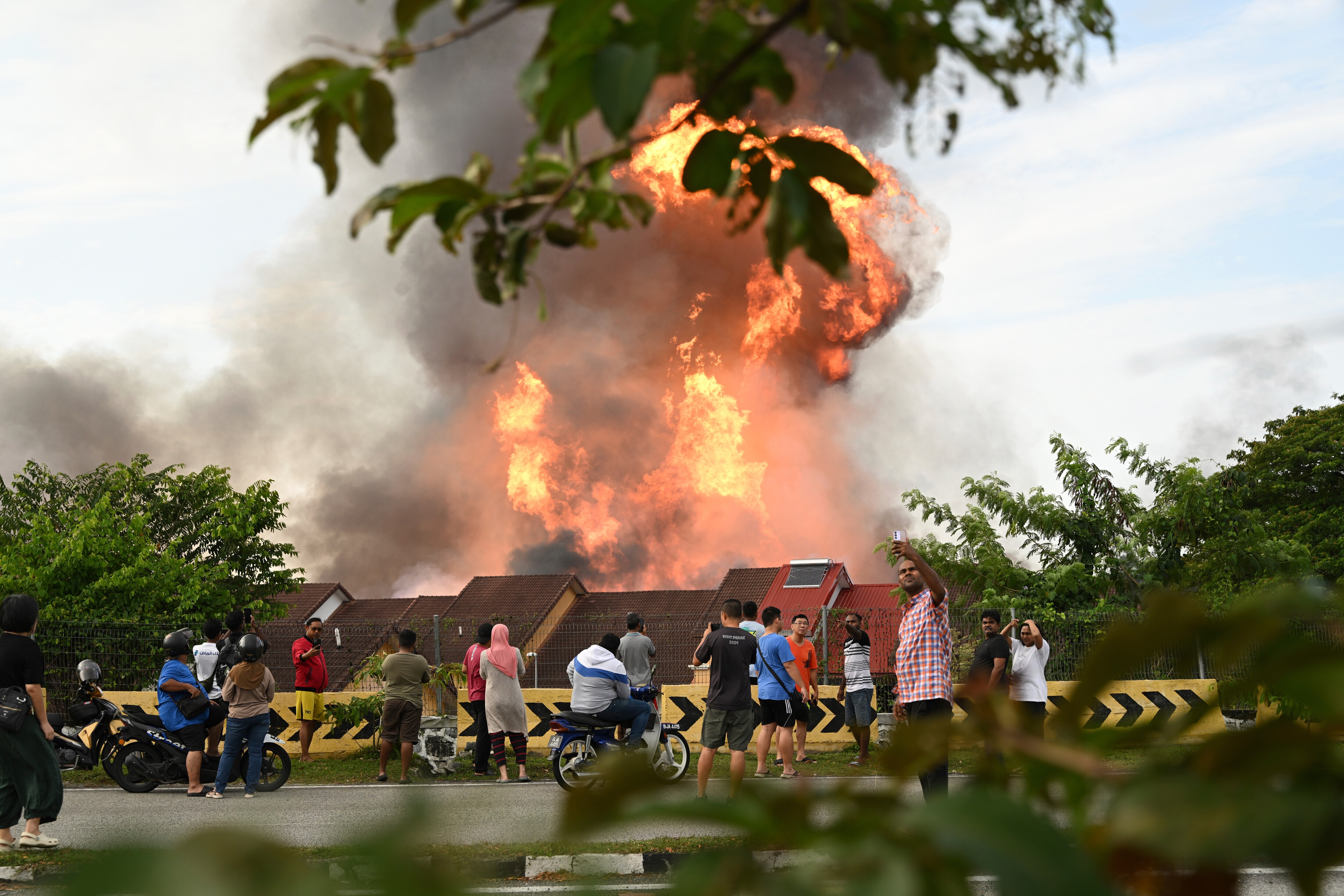 Onlookers standing or sitting on scooters and motorcycles taking photos and watching a large orange fire column above homes