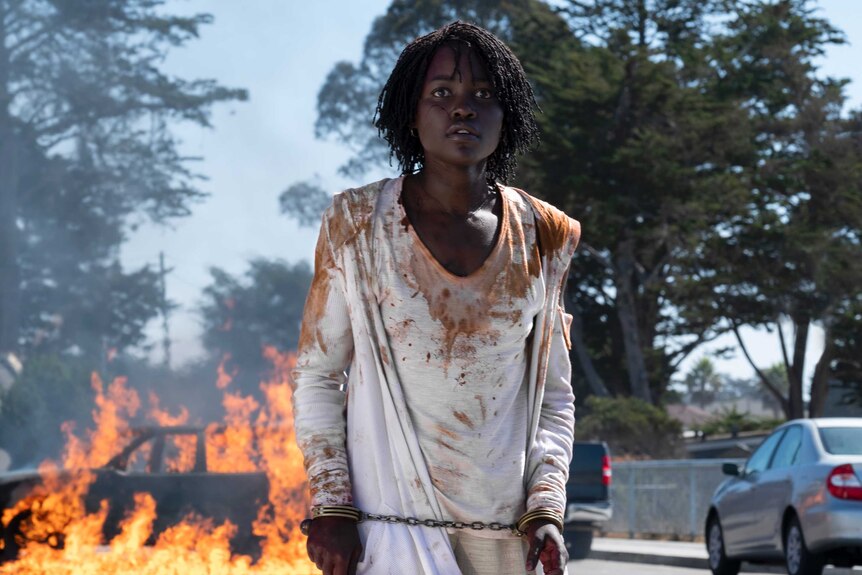 The actor stands, in torn bloody clothes and cuffed, stands in front of a burning car.