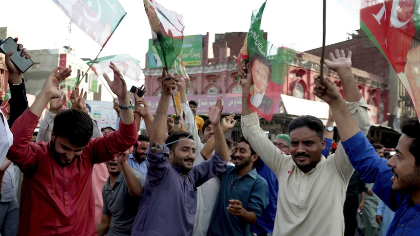 A crowd of Pakistani people are cheering with their hands in the air and holding flags.
