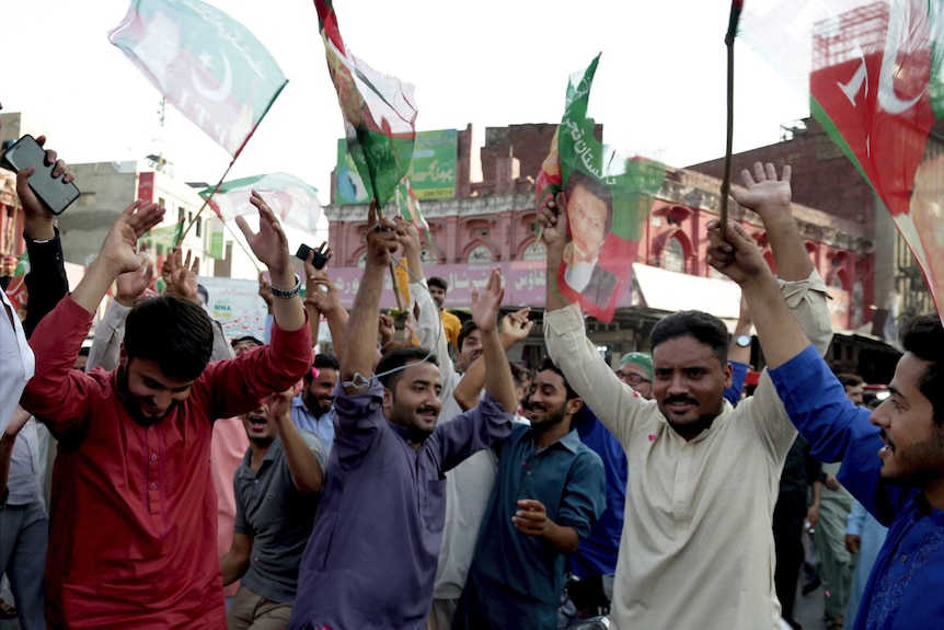 A crowd of Pakistani people are cheering with their hands in the air and holding flags.