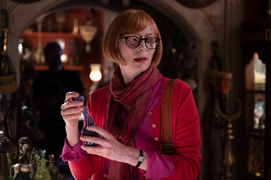 A red-haired woman wearing dark glasses and a red and purple top holds a small blue bottle. 