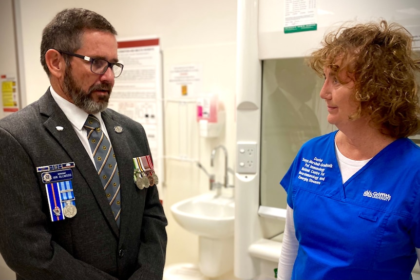 A man in a suit with war medals stands with a health professional.
