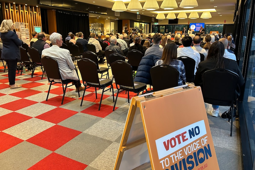 A large group of people in rows of seats listening to a person speaking. An orange sign reads vote no to the voice of division