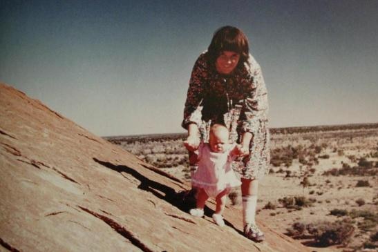 A dark-haired woman holds up an infant by her hands as they stand on the side of Uluru