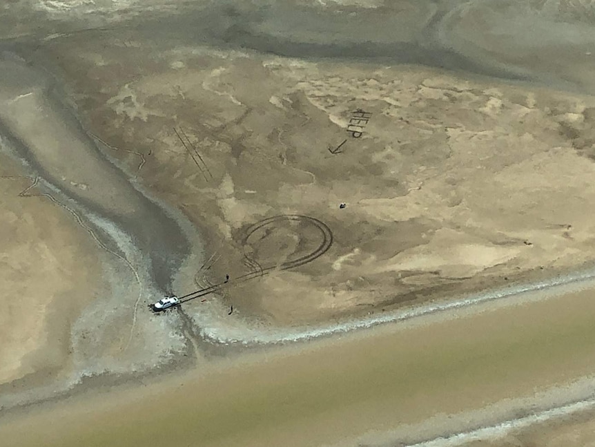 Aerial shot of car bogged in mud and the word "HELP" written in large letters in the sand