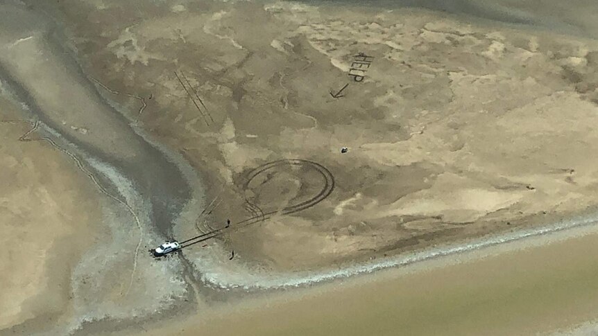 Aerial shot of car bogged in mud and the word "HELP" written in large letters in the sand