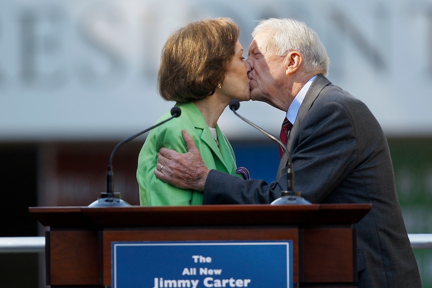 Rosalynn and Jimmy Carter kiss behind a podium.