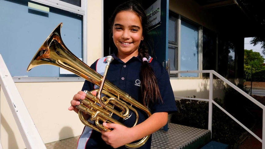 A primary school student stands outside a classroom holding a brass musical instrument: a tenor horn.