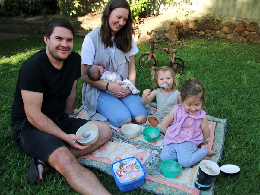 Matt sits on the grass with Rachel, who nurses infant Andie monitors Carter and Billie