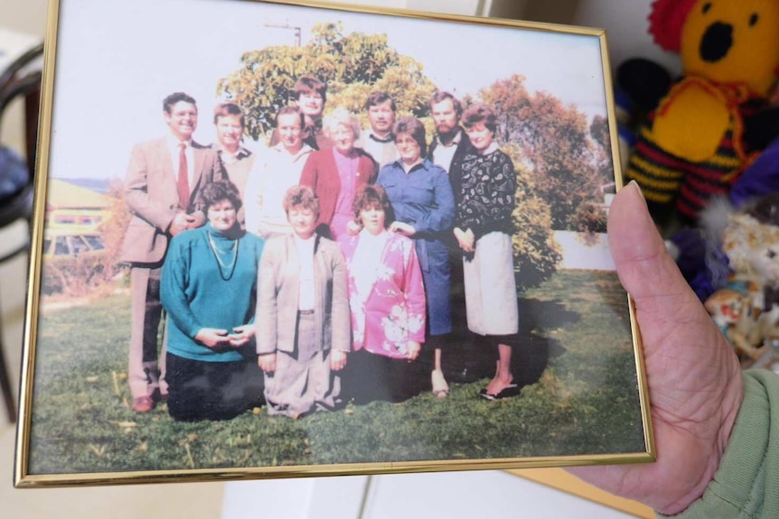 A lady's hand holding a framed photo of a grown-up family pictured in a garden.