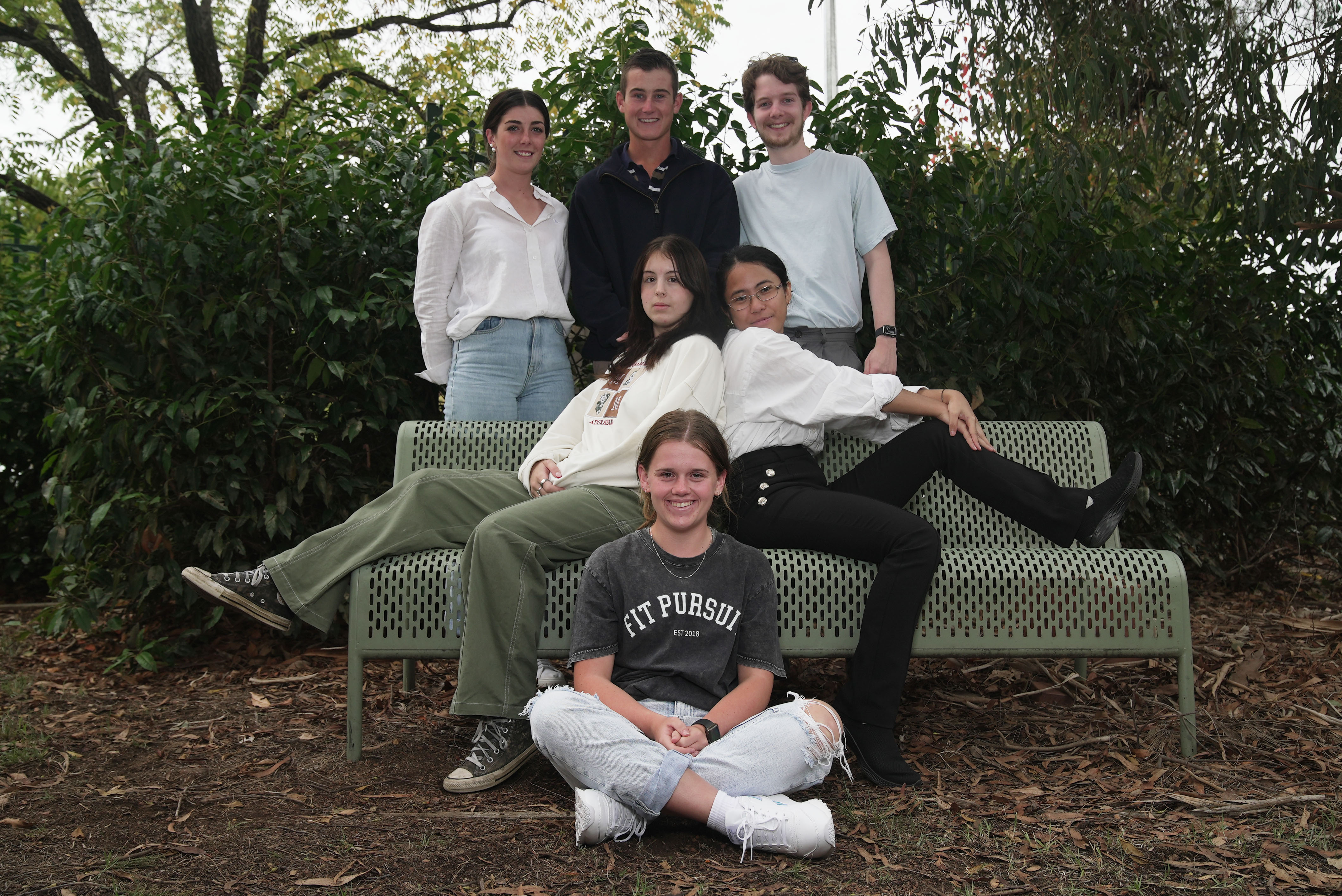 six people, three at the back are standing up smiling, with two people sat on a park bench side by side and one person cross leg