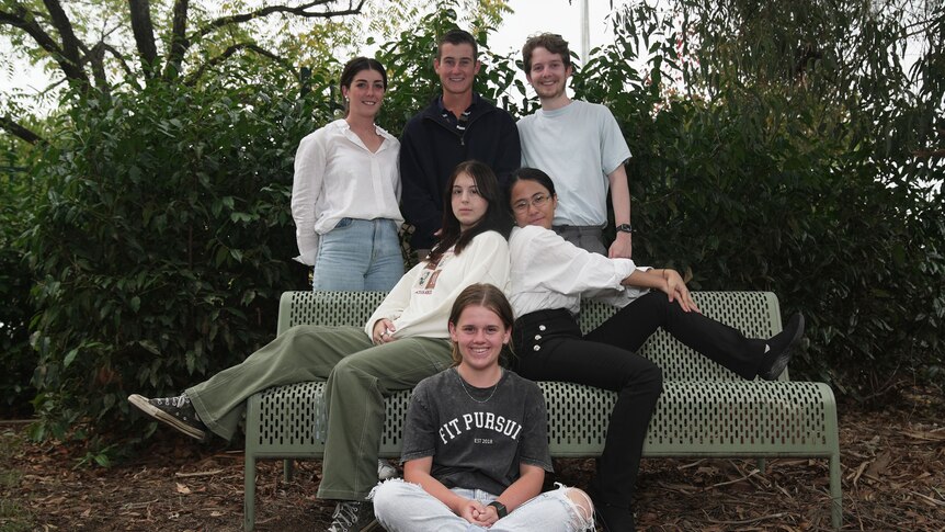 six people, three at the back are standing up smiling, with two people sat on a park bench side by side and one person cross leg