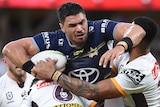 A North Queensland Cowboys NRL player holds the ball with his right arm as he is tackled by two Brisbane Broncos opponents.