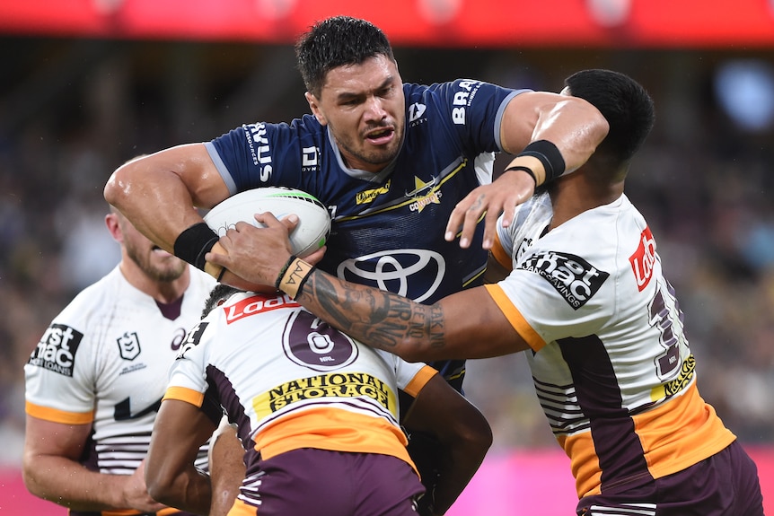 A North Queensland Cowboys NRL player holds the ball with his right arm as he is tackled by two Brisbane Broncos opponents.