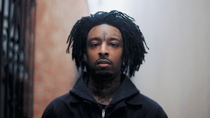 21 Savage with natural hair, a tattooed cross between his eyes, not smiling, headshot, wearing a hoodie