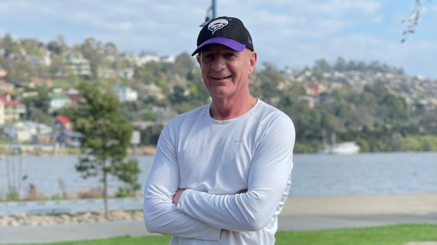 Peter Gutwein is wearing a sports cap and casual clothing and is leaning on a post on the side of a sports oval.