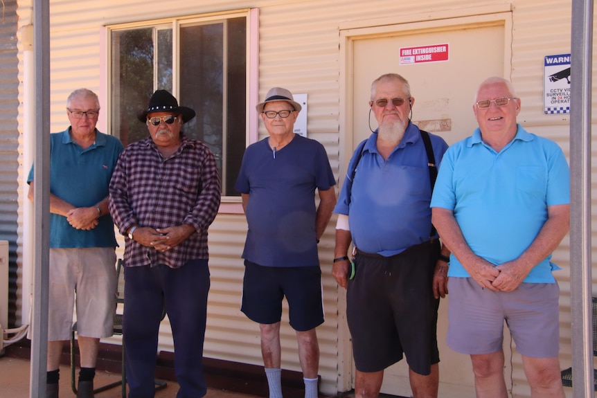 A line of older men standing in front of a corrugated iron building.