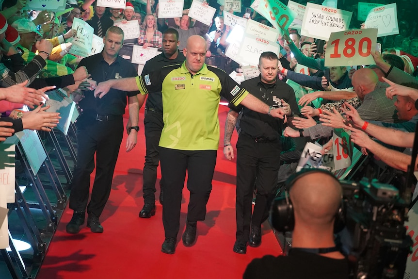 A darts player walks along a red carpet surrounded by security guards, holding his hands out to fist-bump fans.