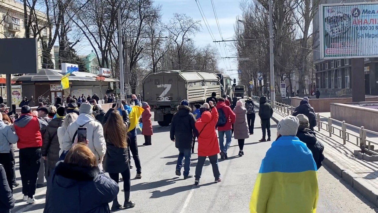 A crowd of people, including some draped in Ukrainian flags, circle a Russian military vehicle.