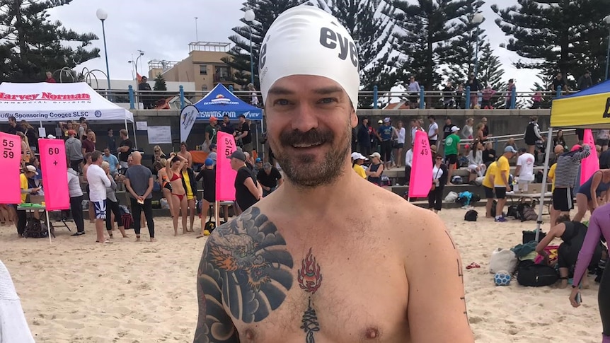 A man wears swimming shorts and a cap and holds goggles on a beach as he prepares for a one kilometre swim.