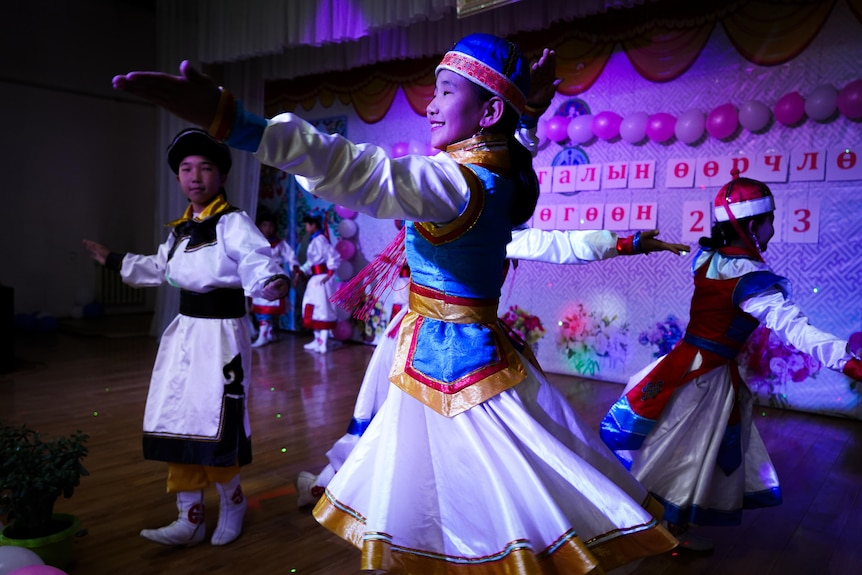 A girl in traditional Mongolian dance costume with bright coat over her dress, and matching hat, smiles while dancing on stage
