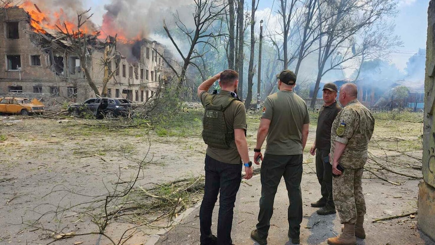 Four men in army gear stand and look at building burning after an airstrike.