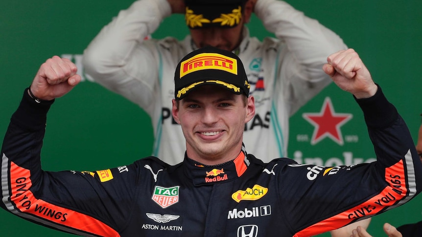 Max Verstappen smiles and holds his hands up as Lewis Hamilton adjusts his hat in the background