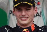 Max Verstappen smiles and holds his hands up as Lewis Hamilton adjusts his hat in the background