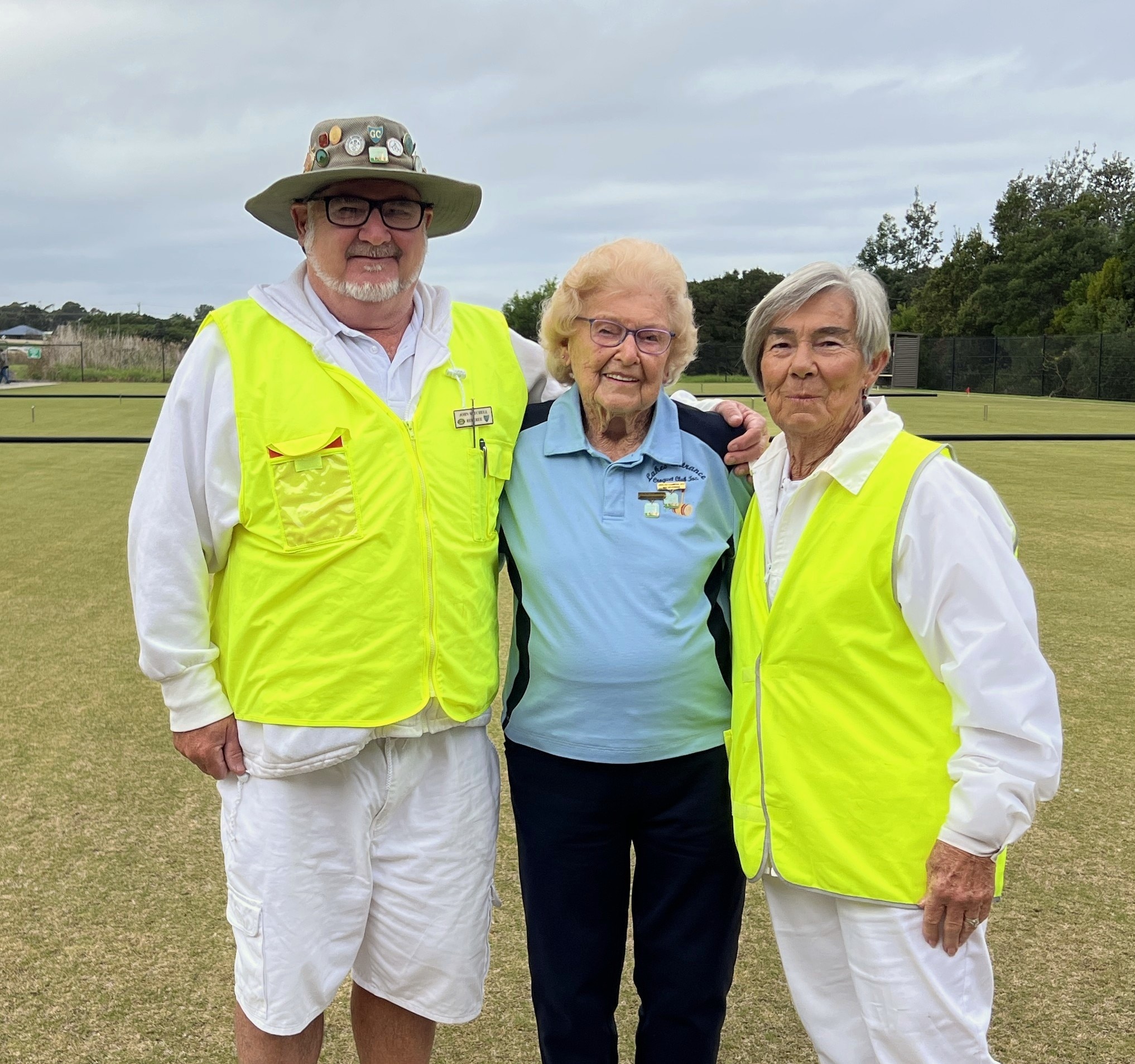A woman wearing a blue polo shirt stands between a man and a woman wearing official vests, on a croquet court. 