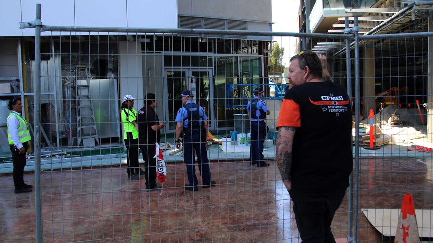 A man leans on a chain link fence at a construction site, while police and security guards speak in the background.