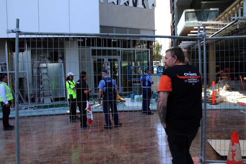 A man leans on a chain link fence at a construction site, while police and security guards speak in the background.