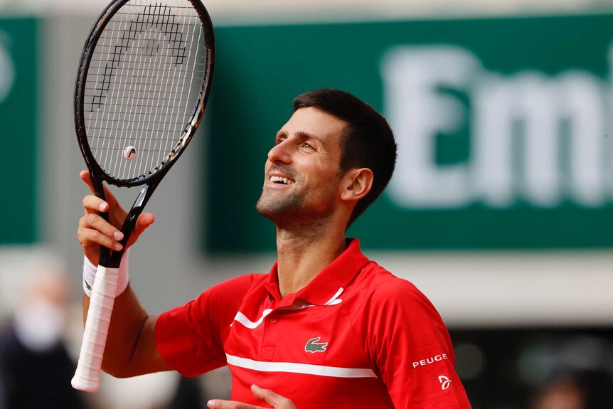 A smiling Novak Djokovic is wearing a red shirt, he looks up and raises his racquet in triumph.