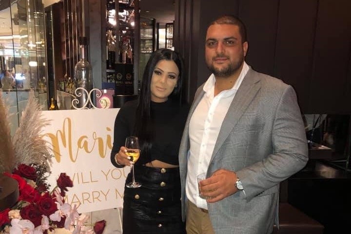 A couple standing together in front of a proposal sign, surrounded by flowers.