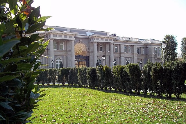 The front entrance to an opulent mansion surrounded by hedges 