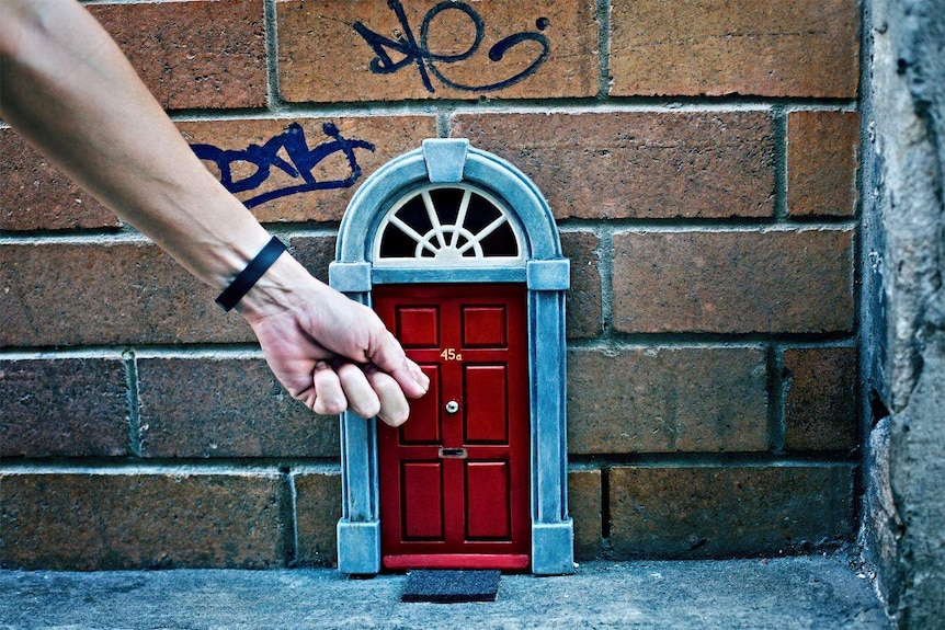 A hand reaches down to knock on a little red door on a wall in Brisbane CBD