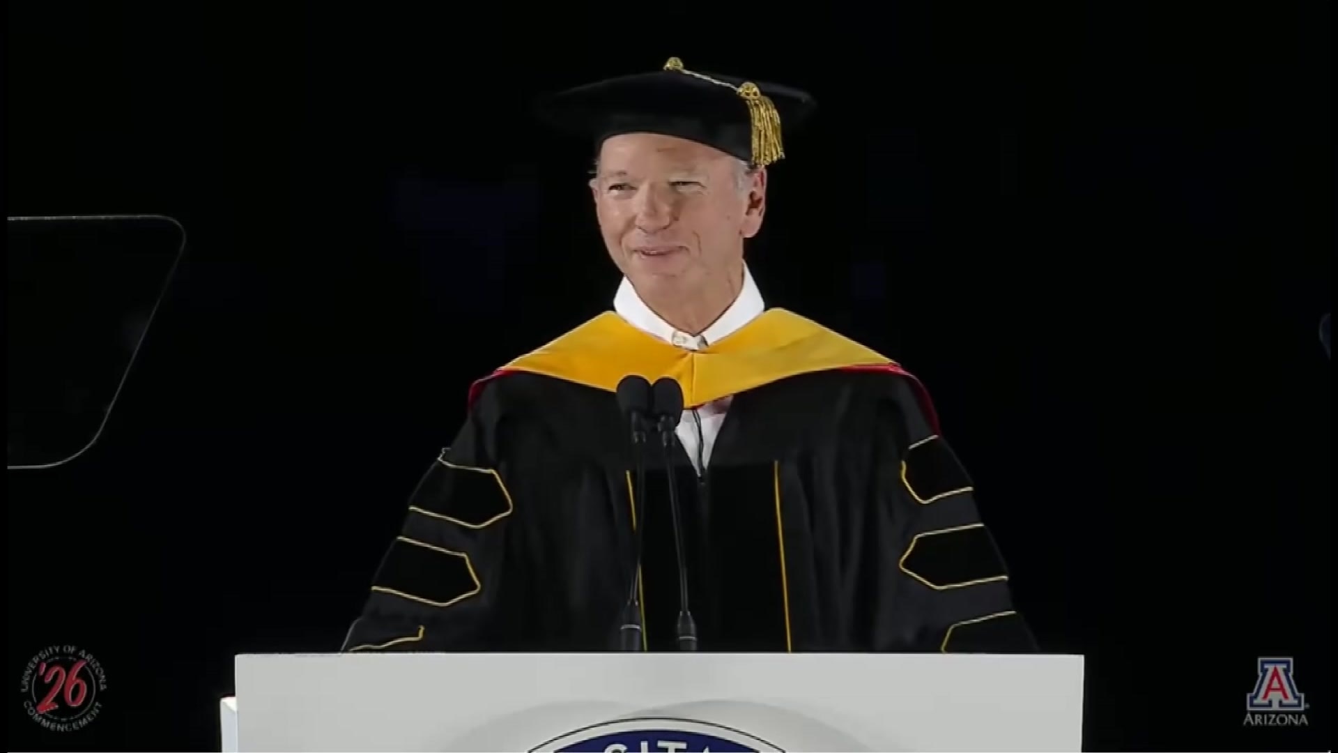 A middle-aged man in a university robe and cap speaks at a podium. 