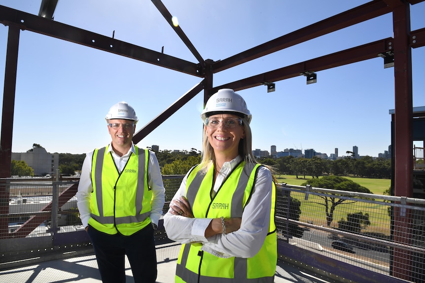A man and woman stand on the top floor of a building under construction, with support beams and a blue sky behind them.