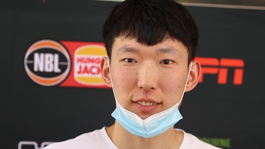 A young Chinese man with a mask pulled down stands at an official event of some kind.